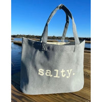 Gray tote bag with 'salty.' text on a wooden dock by a body of water.