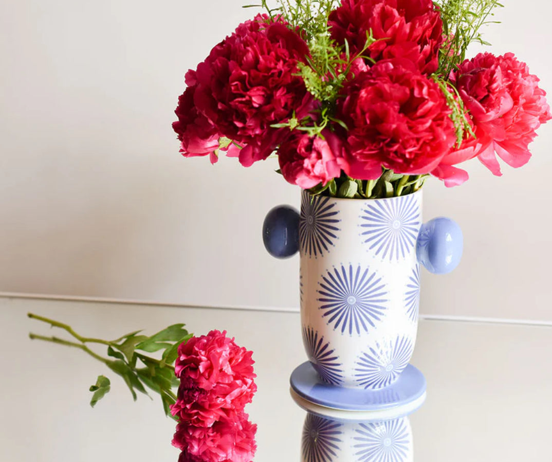 A white vase with blue starburst designs and blue handles filled with red flowers