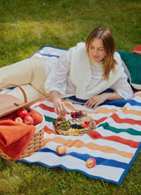 Showing the blanket as a picnic blanket. The blanket has wide stripes alternating with white, orange, green, yellow and blue 