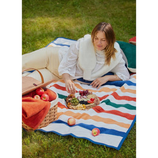 Showing the blanket as a picnic blanket. The blanket has wide stripes alternating with white, orange, green, yellow and blue 