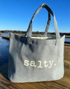 Gray tote bag with 'salty.' text on a wooden dock by a body of water.